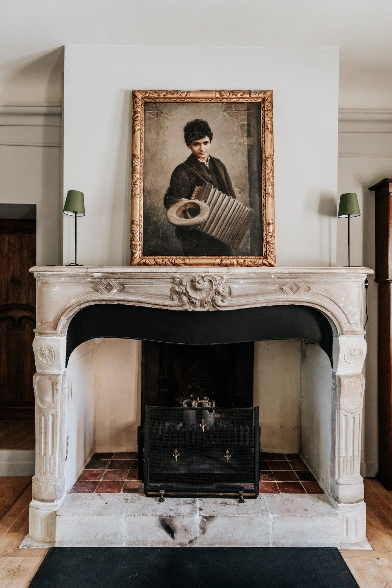 Une peinture de style vintage représentant une personne avec un accordéon orne le mur au-dessus d'une cheminée en pierre classique. La cheminée est dotée d'un manteau orné et d'un écran sombre à l'avant. Deux petites lampes encadrent le portrait sur le manteau.