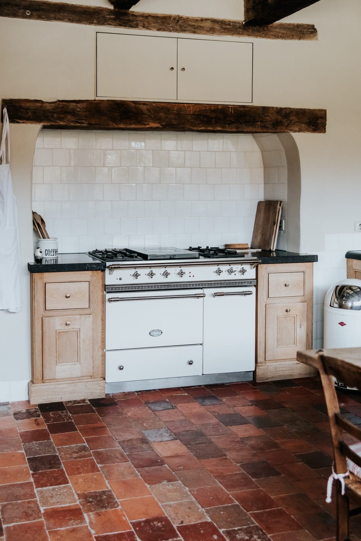 La cuisine rustique est dotée d'une cuisinière de style vintage avec des meubles en bois. Le dosseret est en carrelage blanc et des poutres en bois sont apparentes au-dessus. Le sol est en carrelage rouge et marron. Une théière et une planche à découper sont posées sur le comptoir.