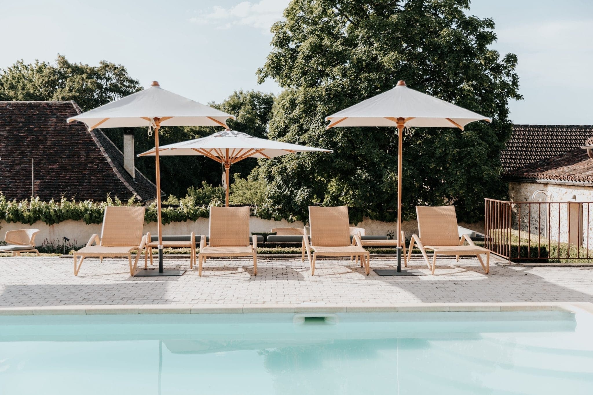 Une scène sereine au bord d'une piscine extérieure avec cinq chaises longues en bois sous de grands parasols. La piscine est au premier plan, et des arbres verts luxuriants et des bâtiments rustiques sont à l'arrière-plan sous un ciel ensoleillé.