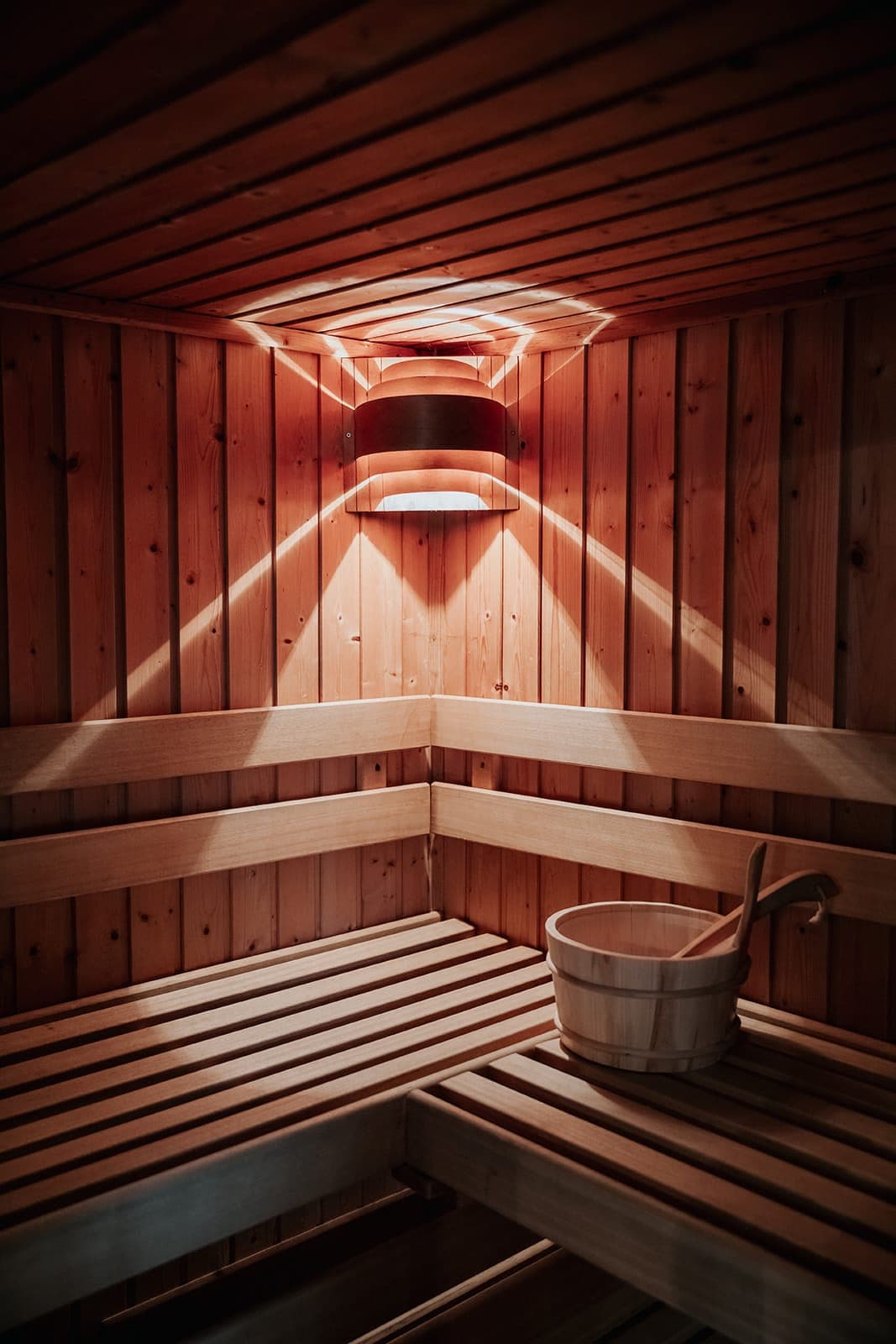 Un intérieur de sauna en bois avec un éclairage chaleureux. Les bancs et les murs sont en bois clair. Un seau en bois avec une louche est posé sur l'un des bancs.