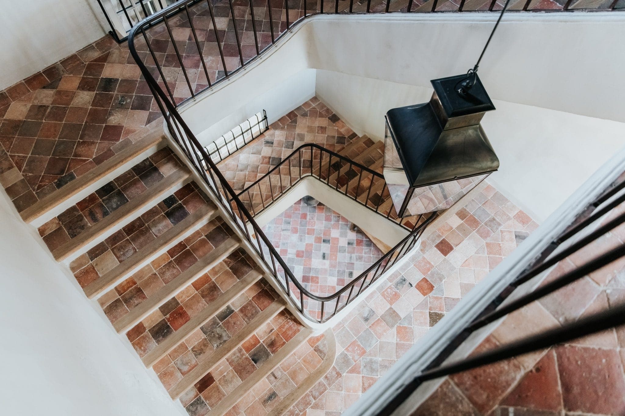 Vue aérienne d'un escalier à plusieurs niveaux avec rampes en fer forgé. Les marches et le sol sont recouverts de carreaux en terre cuite et marron, formant un motif géométrique. Une grande suspension sombre est suspendue au centre au-dessus du sol carrelé en dessous.