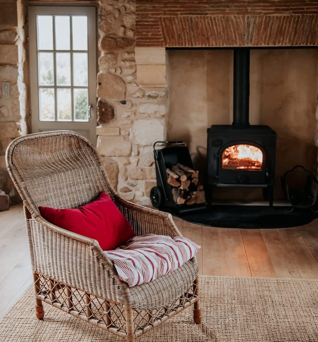 Une pièce cosy avec un fauteuil en osier, orné d'un coussin rouge et d'un oreiller rayé, posé sur un tapis tissé. En arrière-plan, un poêle à bois est encastré dans un mur de pierre et de brique avec des bûches empilées à proximité, créant une ambiance chaleureuse.