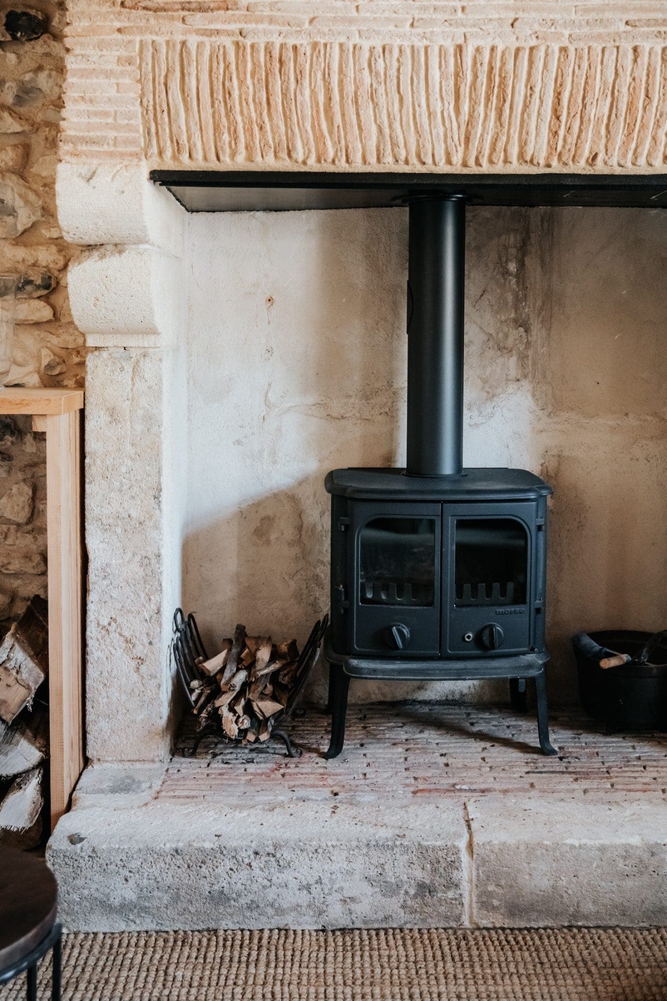 Un poêle à bois rustique est installé devant une cheminée en pierre. Le bois de chauffage est soigneusement empilé d'un côté et un porte-outils en métal noir se trouve de l'autre. La scène dégage une atmosphère chaleureuse et vintage.