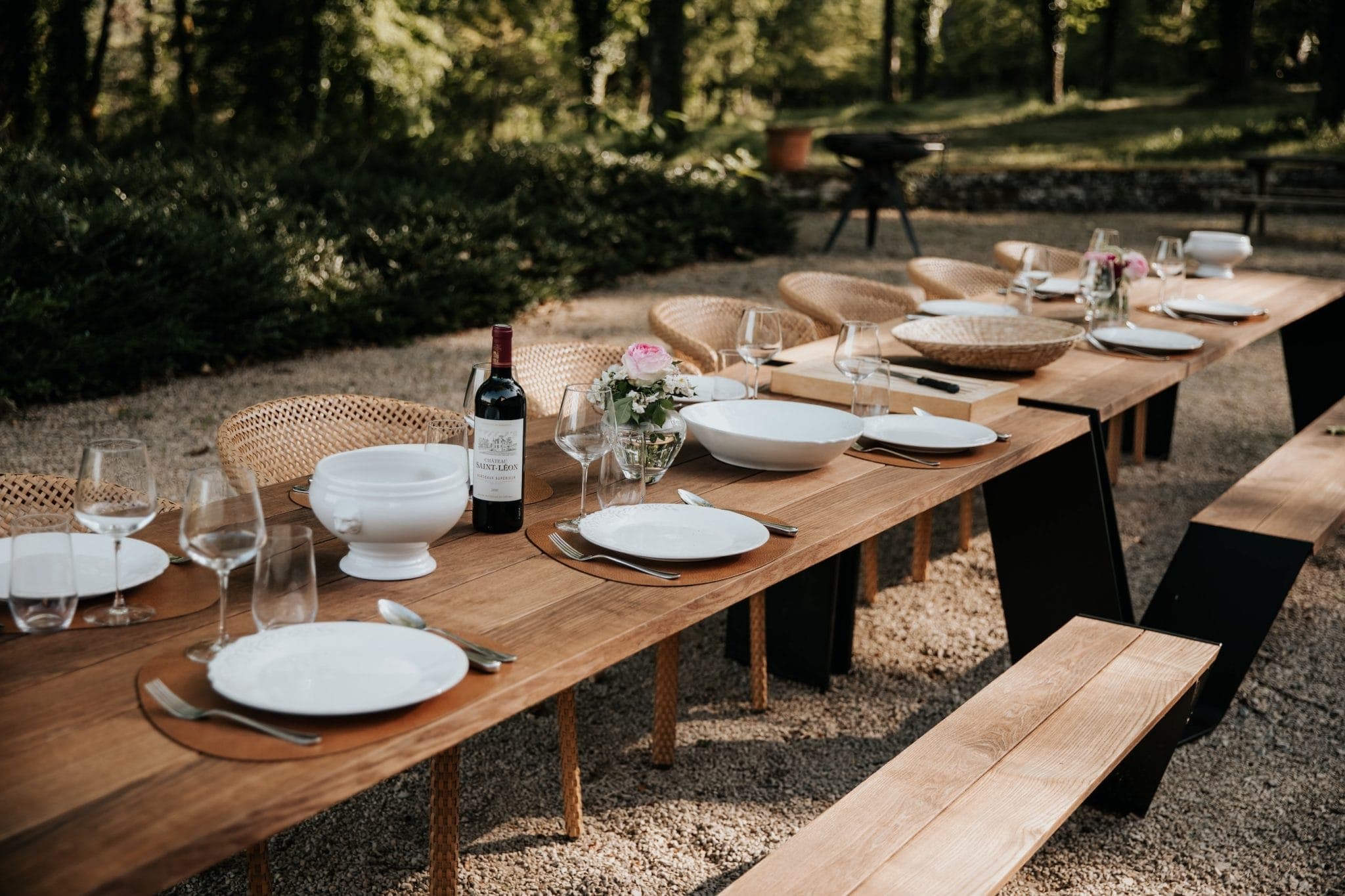 Une longue table d'extérieur en bois dressée pour un repas avec des assiettes blanches, des verres à vin, des couverts, une bouteille de vin rouge et des centres de table floraux. Des bancs en bois bordent chaque côté de la table et des arbres verts forment l'arrière-plan.