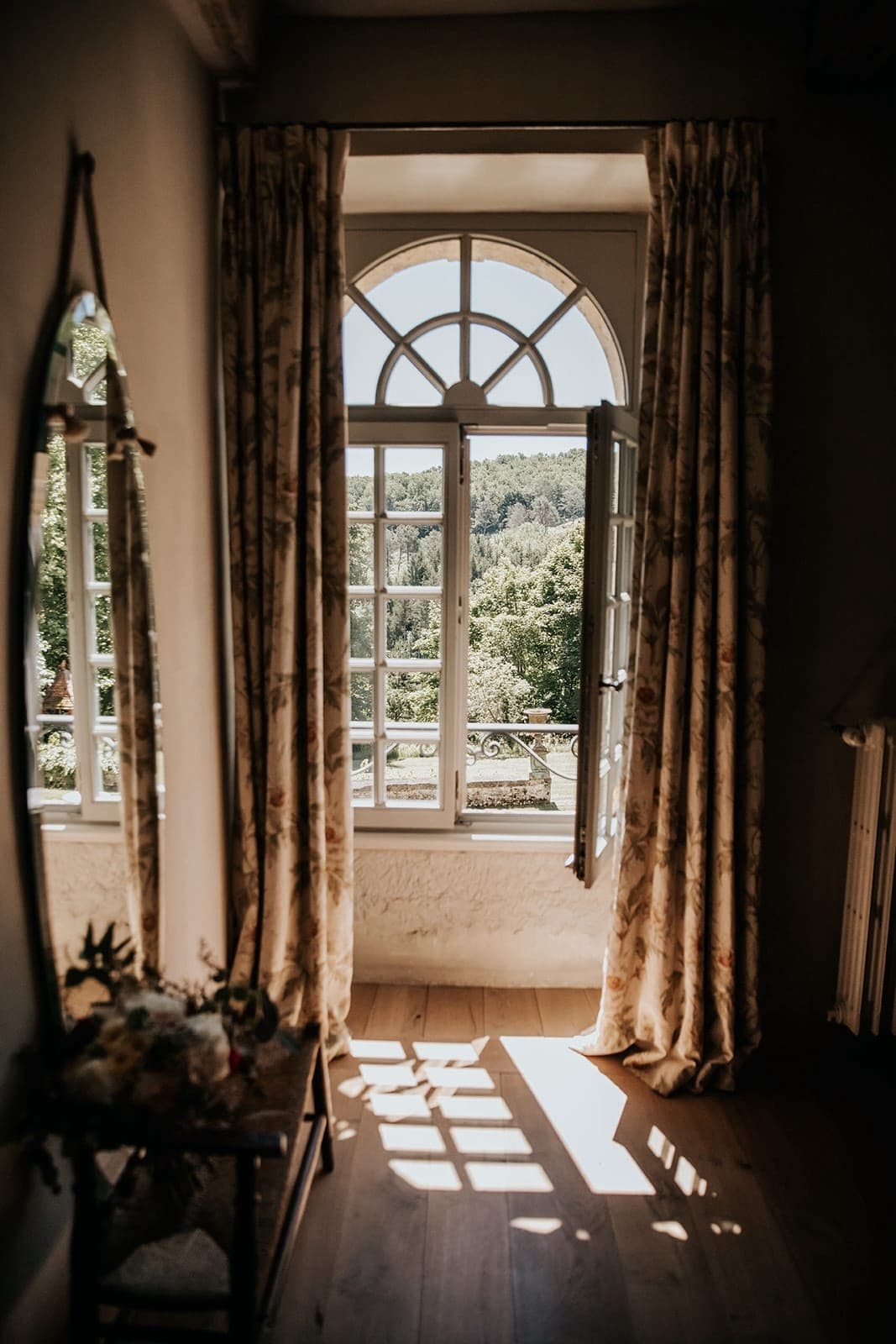 La lumière du soleil pénètre à travers une fenêtre ouverte et cintrée avec des rideaux fleuris, projetant des ombres sur un plancher en bois dans une pièce confortable. Un miroir s'appuie contre le mur, reflétant une partie de la vue sereine sur l'extérieur des arbres et de la verdure.