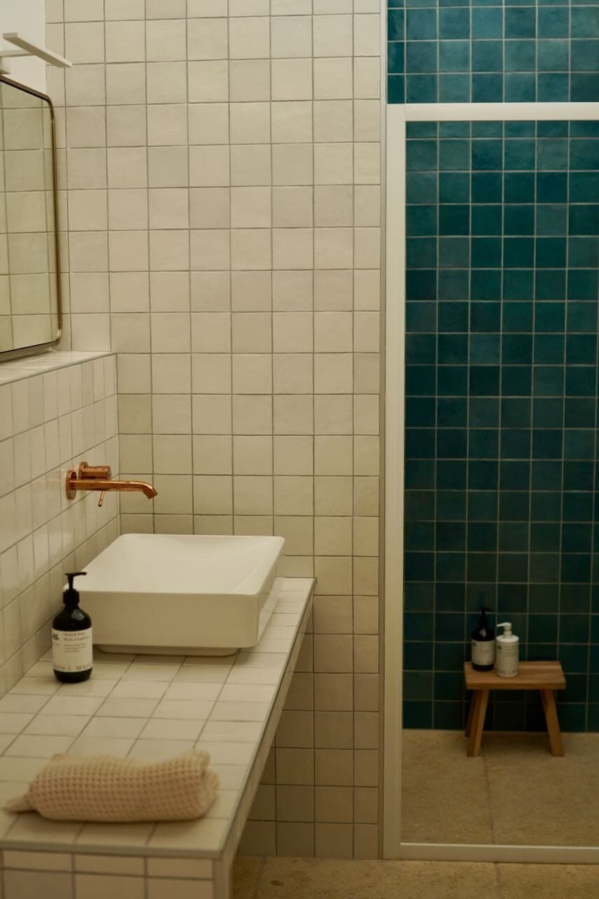 A bathroom has square white tiles and a white countertop with a rectangular sink and copper faucet. There are bottles on the counter and a rolled towel. A shower with teal tiles and a wooden stool with bottles on it is visible.