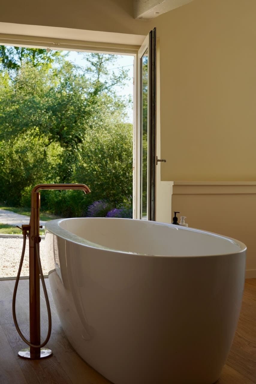 A freestanding bathtub with copper faucet is positioned near an open window, offering a view of the lush greenery outside. Sunlight fills the room, underlining the warm, welcoming atmosphere of the bathroom.