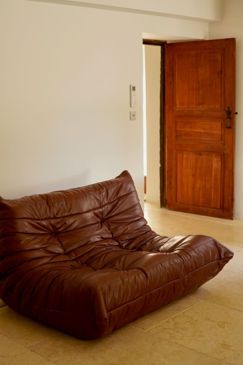 A brown leather sofa rests on a light-colored floor in a minimalist room. The background is composed of a plain off-white wall and an open wooden door, creating a warm, welcoming atmosphere.