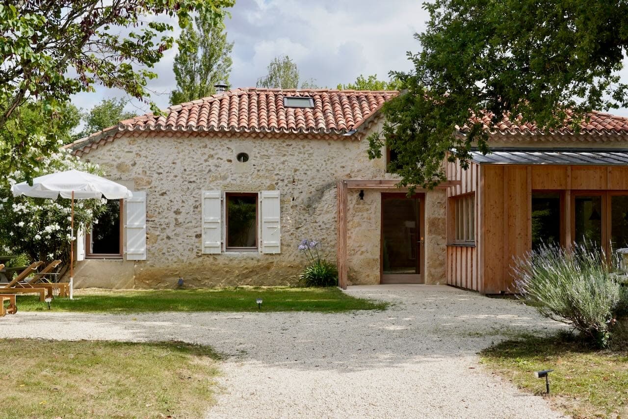 Charming stone house with red-tiled roof and wooden extension surrounded by greenery. The facade features white shutters and a gravel driveway, with a patio shaded by a white parasol and outdoor chairs on the lawn.