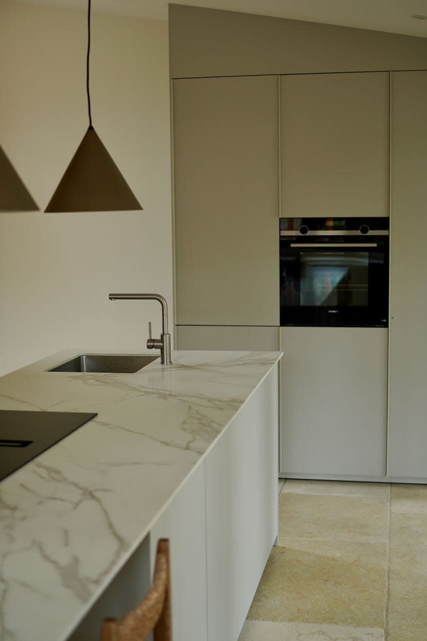 A modern kitchen with elegant marble worktop, integrated sink and stove. Minimalist pendant lights hang above. The background features built-in cabinets with an oven, creating a sleek, contemporary design.