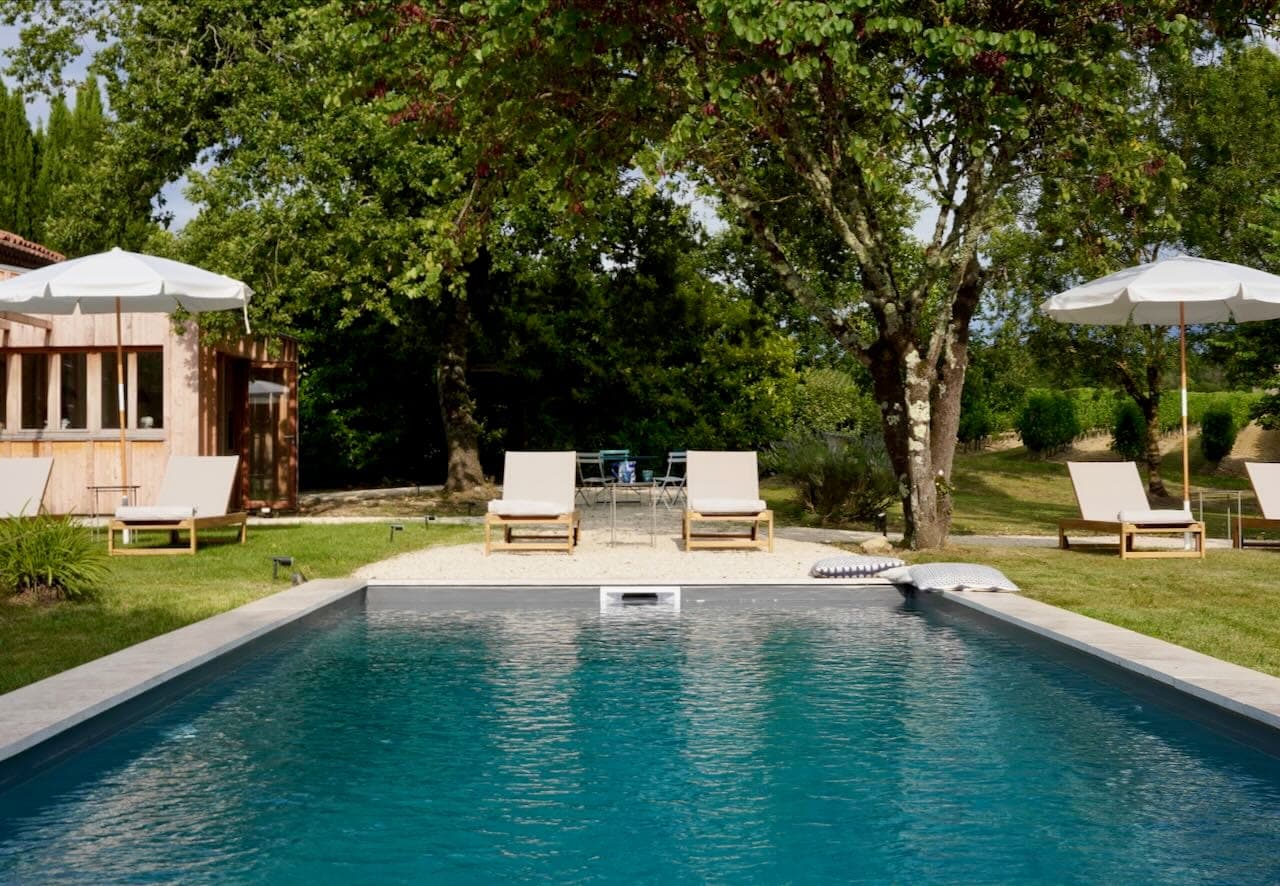 A serene outdoor scene with a rectangular pool surrounded by lush vegetation. Sun loungers and parasols are neatly arranged around the poolside, and a wooden building is visible to the side, all under a clear blue sky.