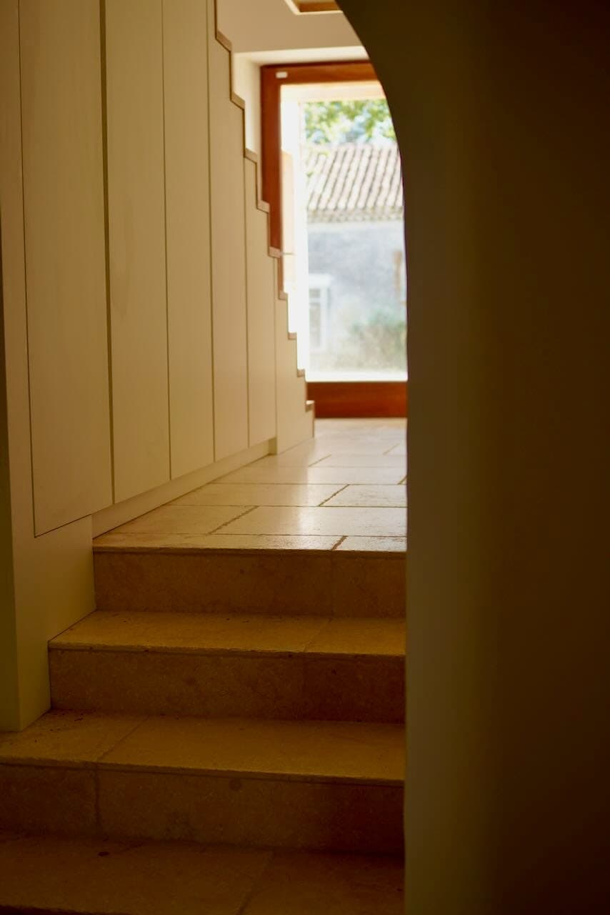 A narrow, dimly-lit staircase, with stone steps leading up to a wooden door. Sunlight filters through the glass, illuminating part of the walls and floor. A small part of a window with greenery outside is visible.