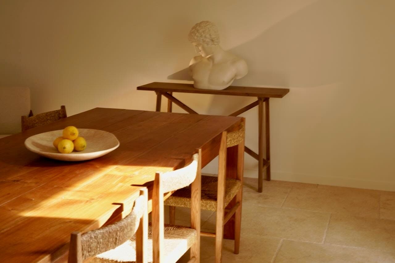 Une salle à manger chaleureuse et ensoleillée est dotée d'une table et de chaises en bois. Un bol de pommes jaunes est posé sur la table. À l'arrière-plan, un buste classique repose sur une console contre un mur de couleur claire.