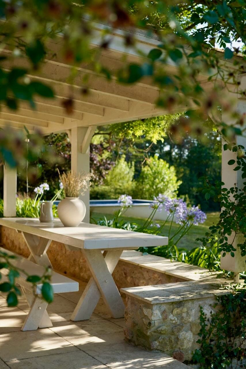 A serene patio scene with a white wooden table with a large vase and pitcher, under a pergola. The space is surrounded by lush greenery and flowers, with sunlight filtering through the leaves. A stone wall borders the patio.