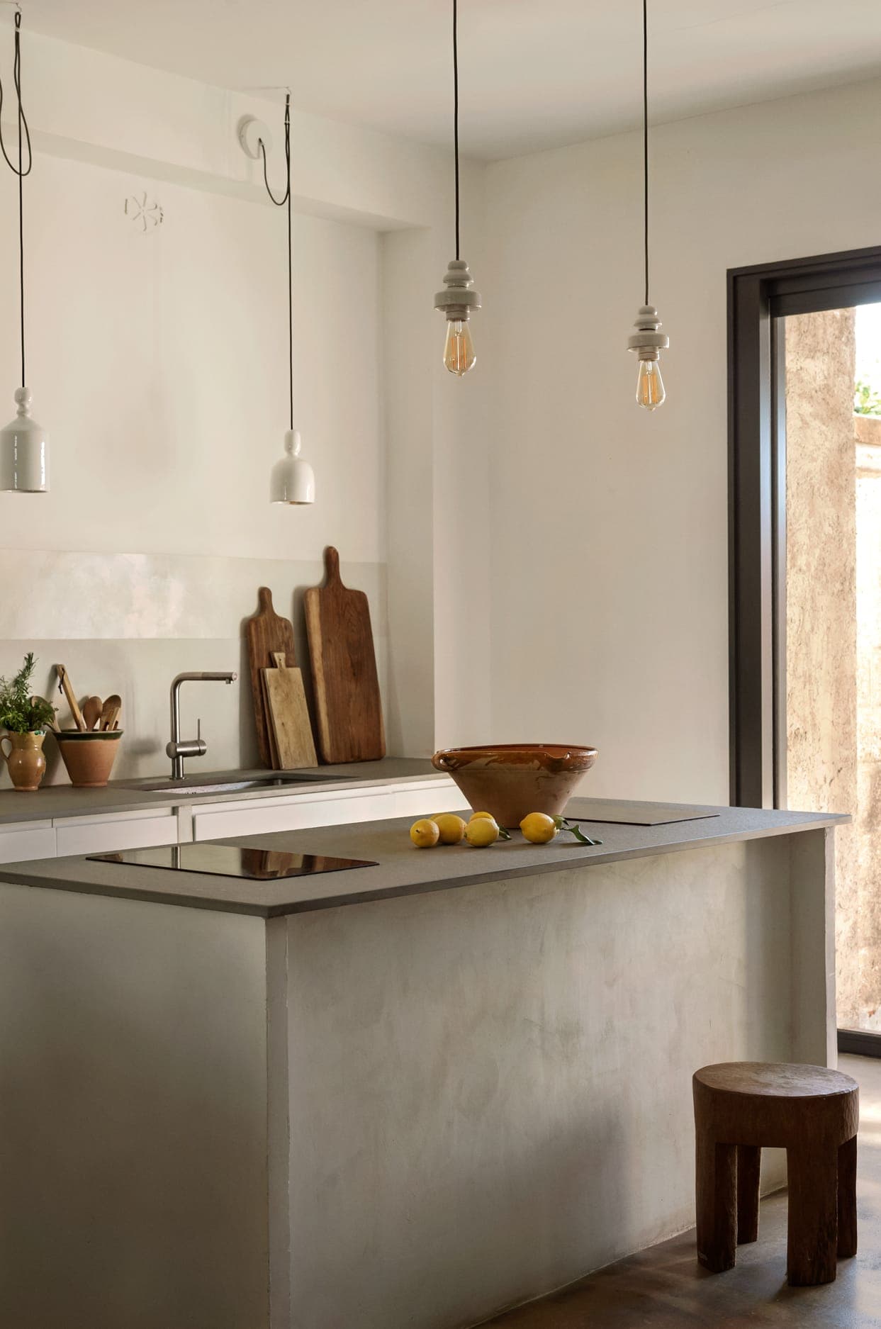 A minimalist kitchen features a concrete island with lemons and a brown bowl on top. Wooden cutting boards stand upright near the sink. Three hanging light bulbs provide illumination. A small wooden stool is placed beside the island. A large window offers natural light.