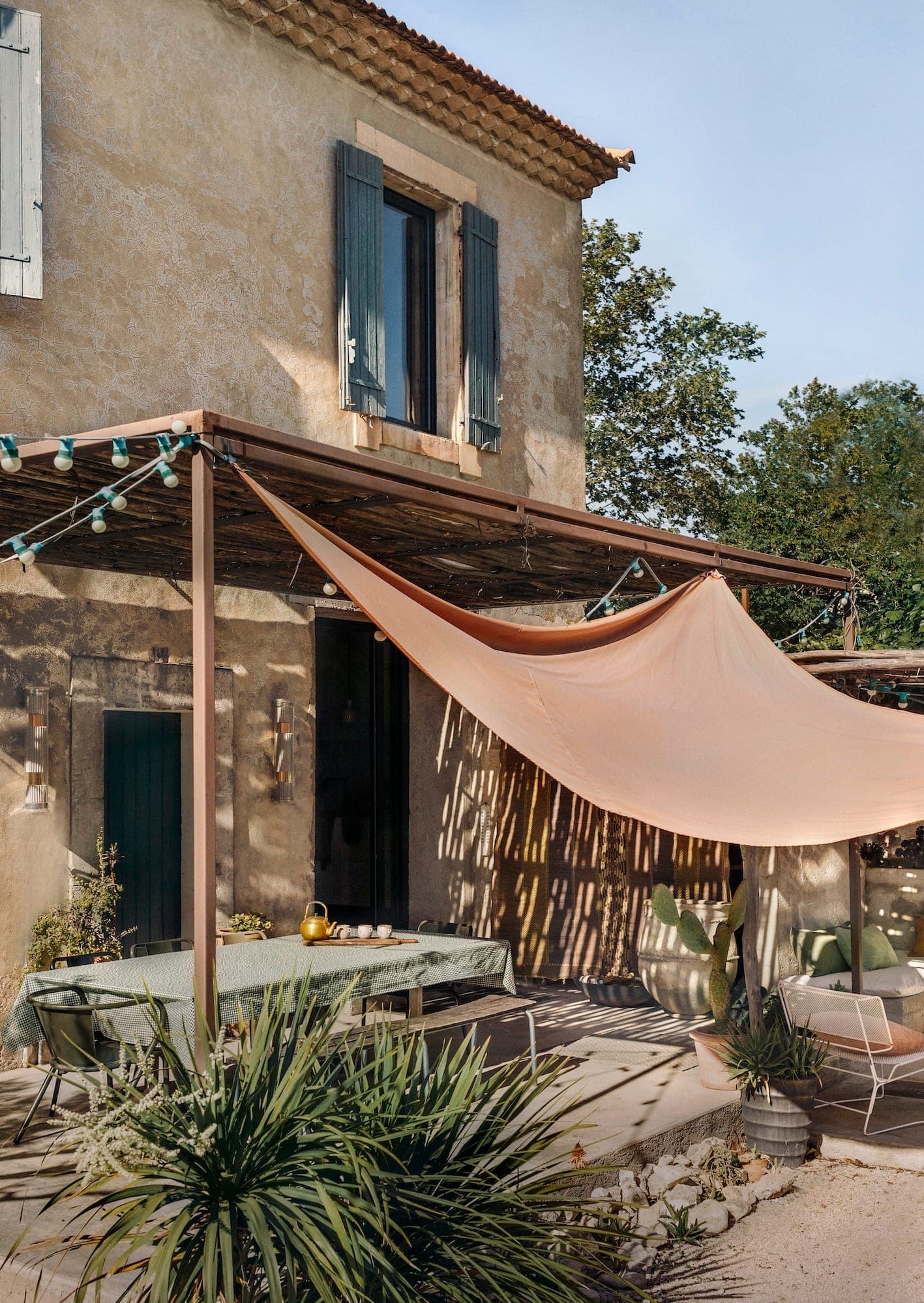 Une maison rustique à deux étages avec des murs en pierre et des volets bleus dispose d'un patio avec une pergola. Un grand store en tissu beige est drapé sur un coin salon avec des chaises vertes et une table. Des plantes luxuriantes décorent le patio.