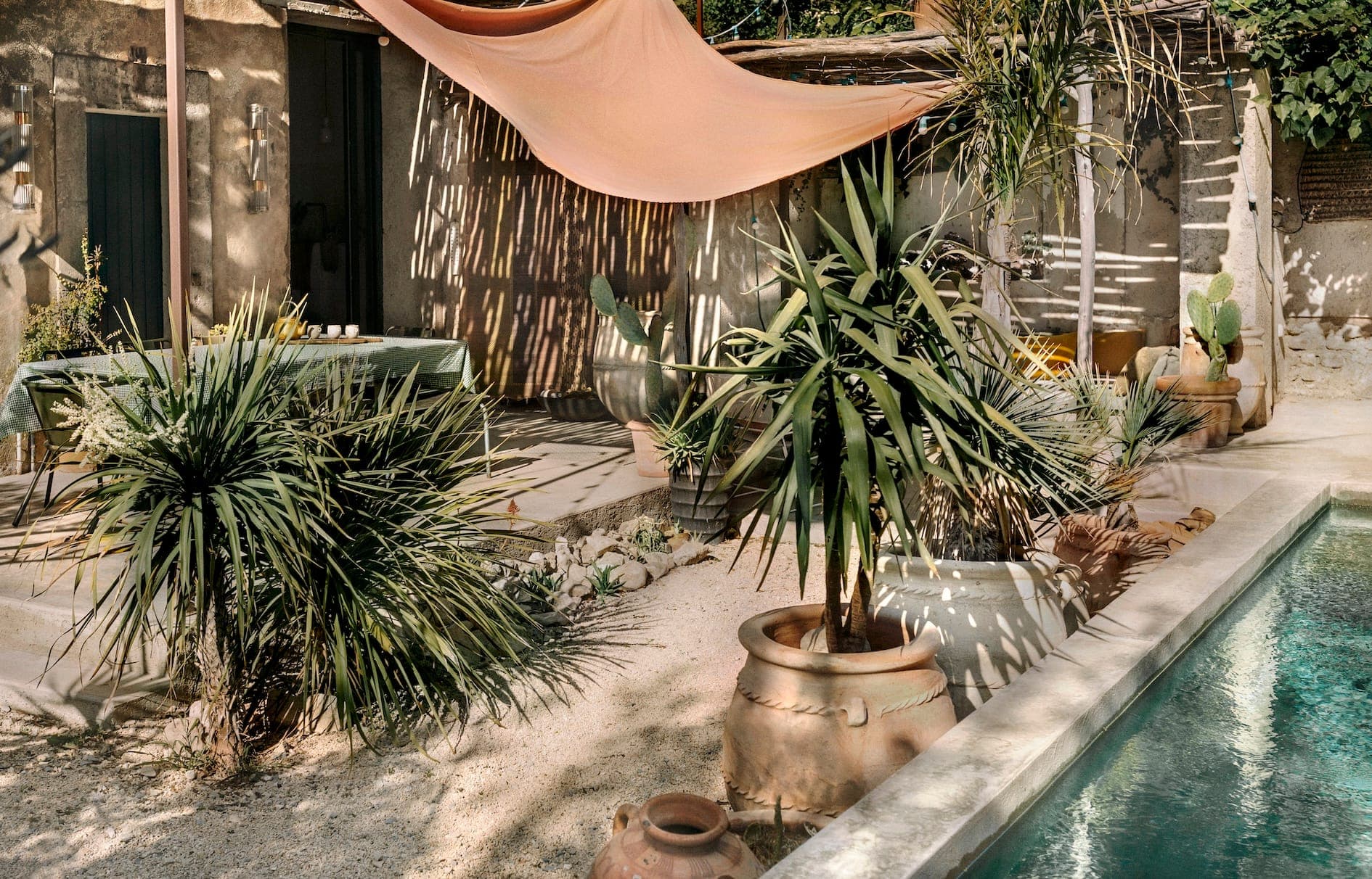 Sunlit patio with lush potted plants beside a turquoise pool. A large salmon-colored fabric canopy provides shade over a rustic dining area. The setting features wooden textures and green foliage, creating a serene, tropical ambiance.