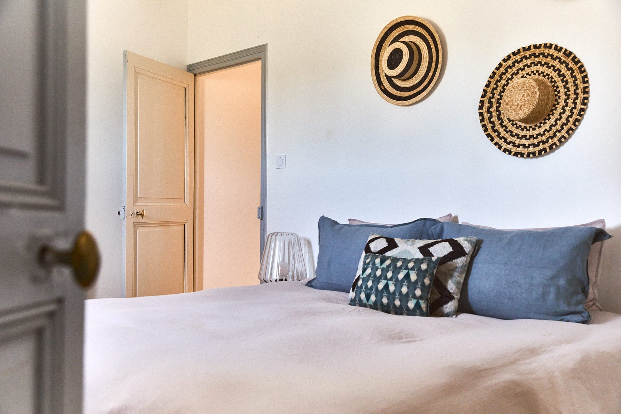 Chambre confortable avec un lit bien fait avec des oreillers bleus et un coussin à motifs. Deux paniers tressés décoratifs sont accrochés au mur blanc au-dessus du lit. Une porte est partiellement ouverte, révélant un couloir doucement éclairé.