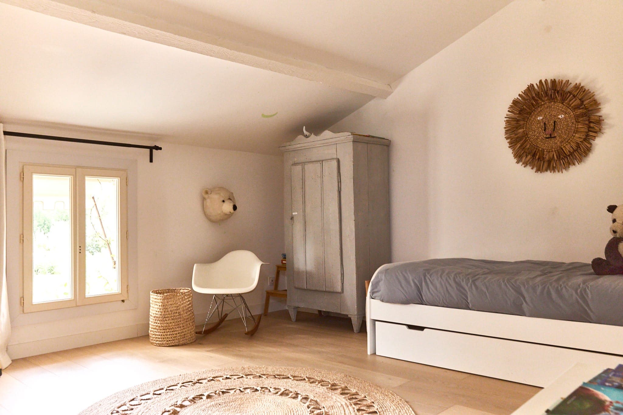 Une chambre confortable avec un lit blanc, une armoire grise et une décoration en osier. Une tête d'ours en peluche est accrochée au mur. La lumière naturelle entre par une fenêtre. Le plancher en bois est partiellement recouvert d'un tapis rond tissé, et il y a une petite chaise blanche et un panier.