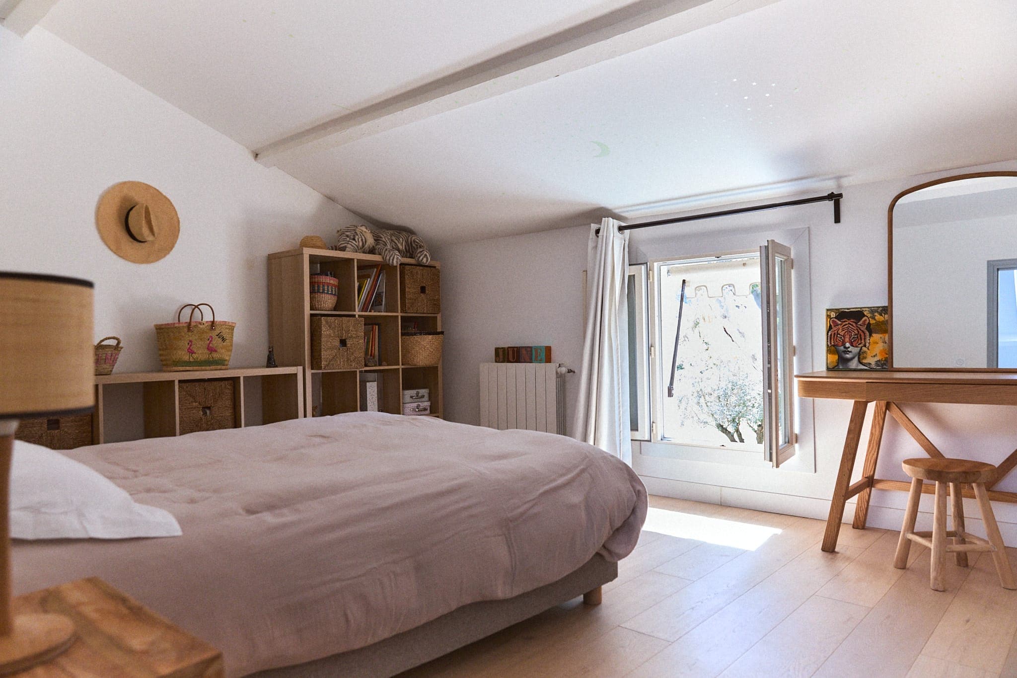 Une chambre lumineuse et minimaliste avec un grand lit, un parquet en bois clair et une fenêtre laissant entrer la lumière naturelle. La pièce comprend un bureau en bois avec un tabouret, un grand miroir, un chapeau et des paniers sur des étagères pour le rangement et la décoration.