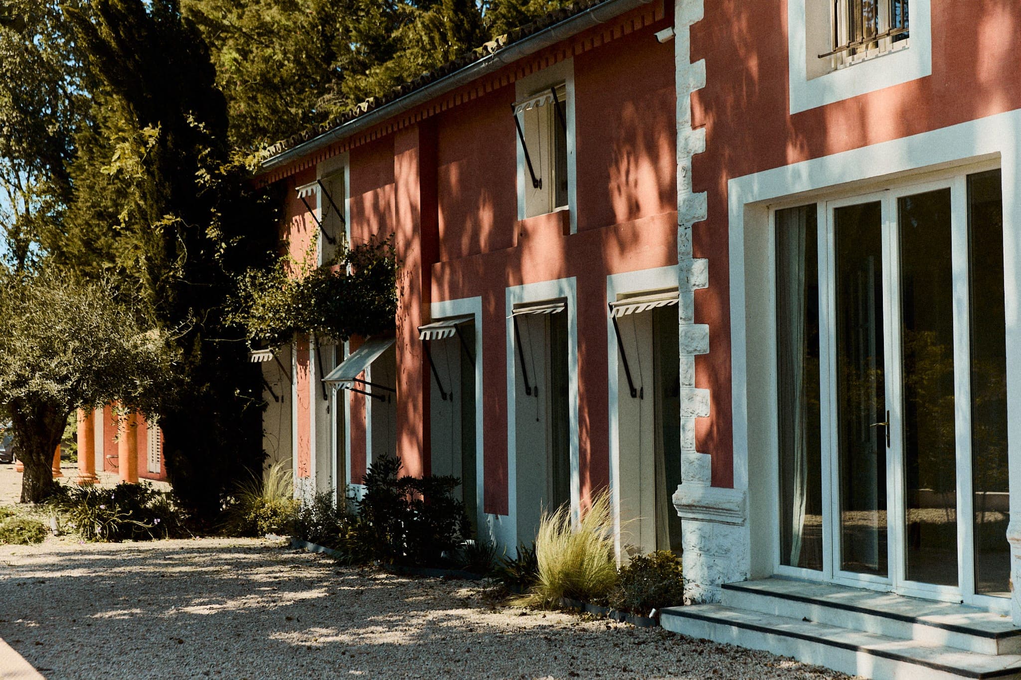 Un bâtiment rouge rustique avec de nombreuses petites fenêtres et portes est entouré d'arbres. L'entrée est dotée de grandes portes vitrées et un chemin de gravier mène au bâtiment, projetant des ombres provenant des arbres voisins.