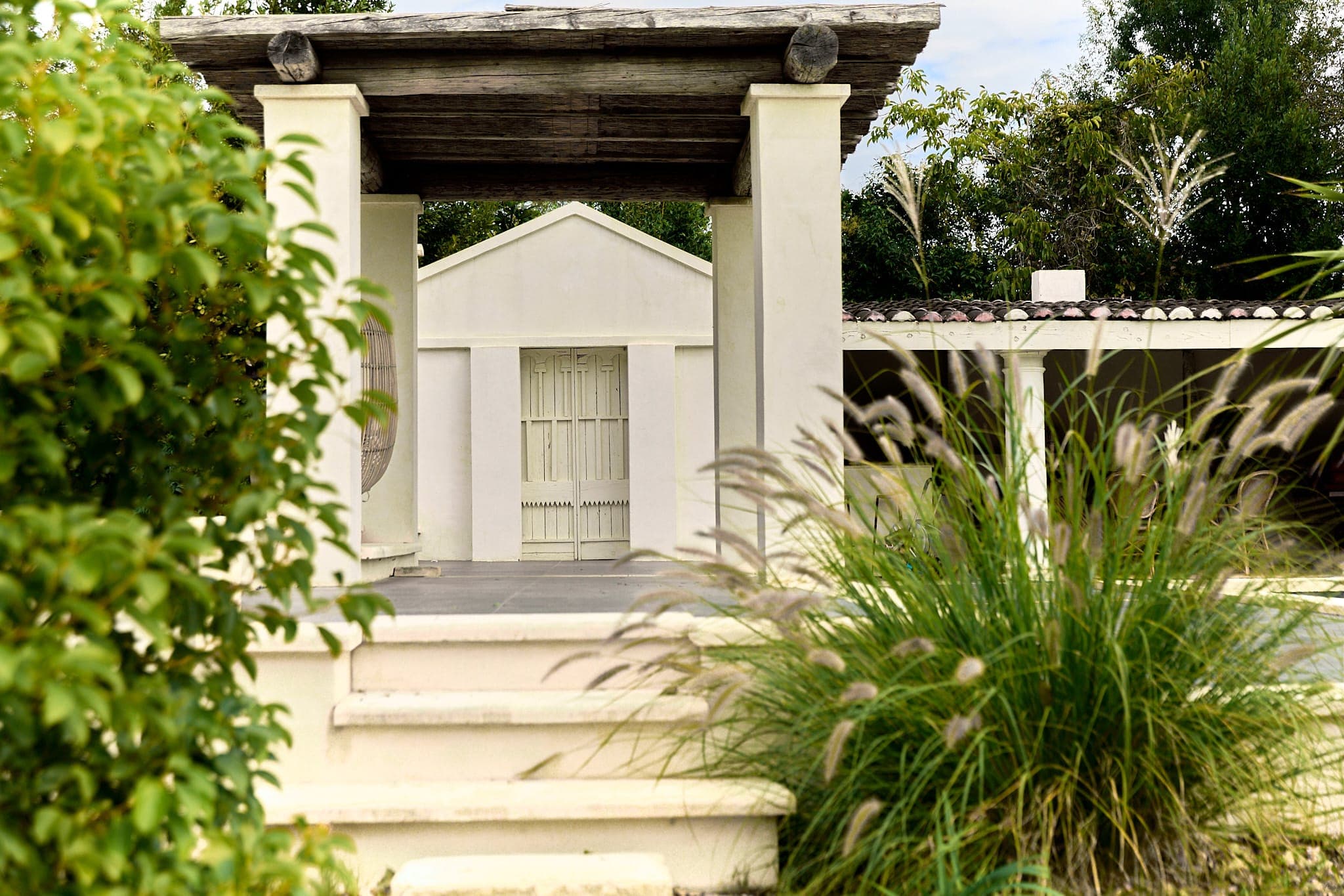 Vue sereine d'une structure blanche à colonnes avec un toit en bois, entourée d'une végétation luxuriante et de graminées ornementales. La scène transmet un sentiment de tranquillité et de beauté naturelle.