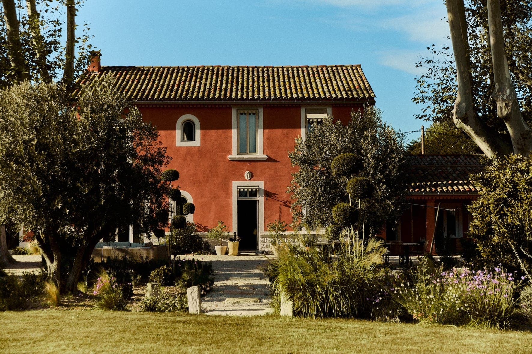 A rustic red house with a tiled roof, surrounded by lush vegetation and trees, and a garden with flowers. The entrance features a pathway with potted plants, under a blue sky with scattered clouds.