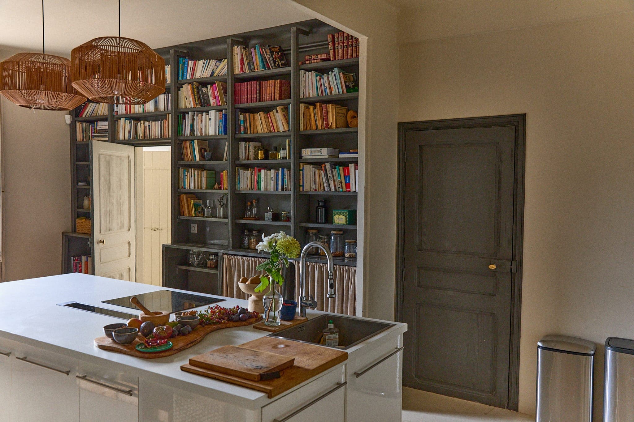 Une cuisine chaleureuse avec un grand îlot avec des planches à découper et des ingrédients frais. Les murs sont dotés d'étagères intégrées remplies de livres. Des lampes suspendues en osier ajoutent de la chaleur, et il y a des portes blanches et une porte grise en arrière-plan.