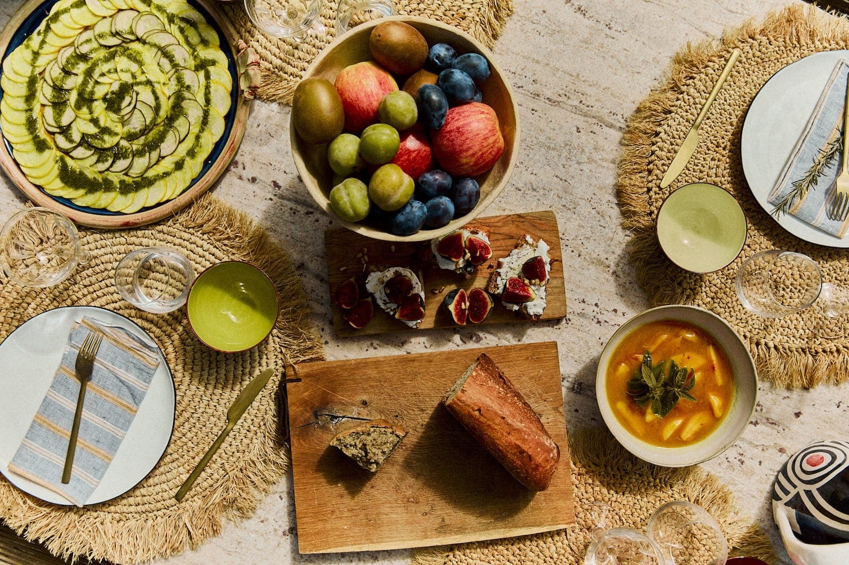 Une table dressée avec des mets variés : une tarte aux courgettes, un bol de fruits mélangés, du pain tranché, des petites tartelettes aux fruits et un bol de dessert garni de menthe. La table est dressée avec des sets de table tressés et de la vaisselle élégante.