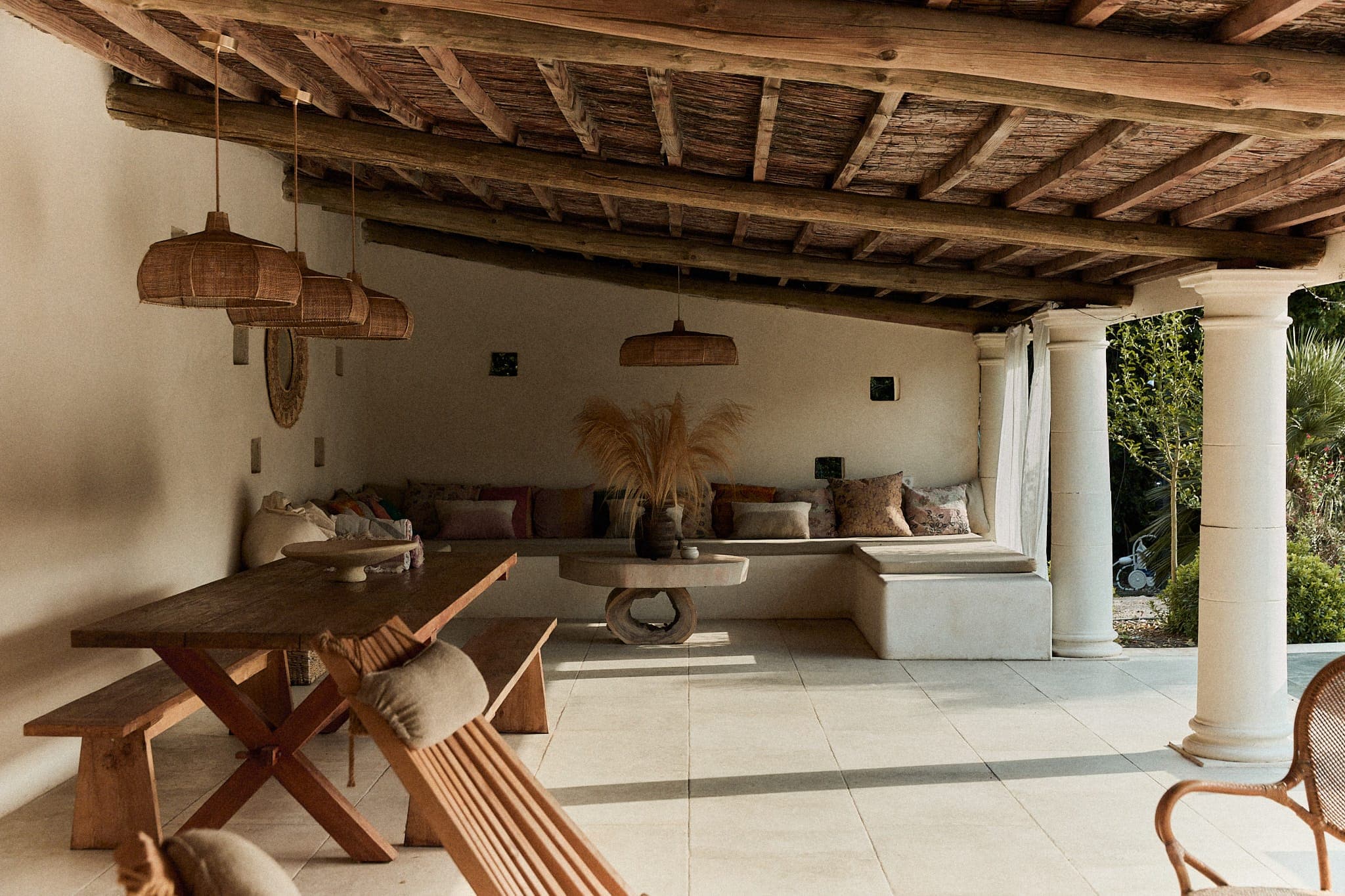 Une terrasse extérieure rustique comprend une table et des chaises en bois, des suspensions en osier et des sièges rembourrés le long du mur. Un plafond à poutres en bois et des colonnes blanches encadrent l'espace. Des plantes séchées et des coussins moelleux ajoutent une touche chaleureuse.