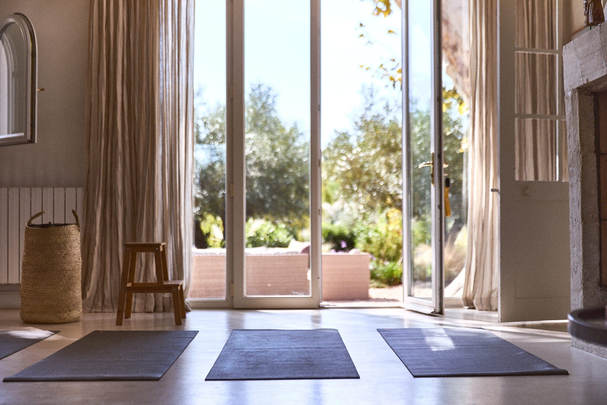 Un espace de yoga intérieur serein avec trois tapis au sol, bordé d'un panier en osier et d'un petit tabouret en bois. La lumière du soleil filtre à travers de grandes portes vitrées, ouvrant sur un jardin luxuriant avec des arbres et de la verdure. Des rideaux couleur crème pendent de chaque côté.