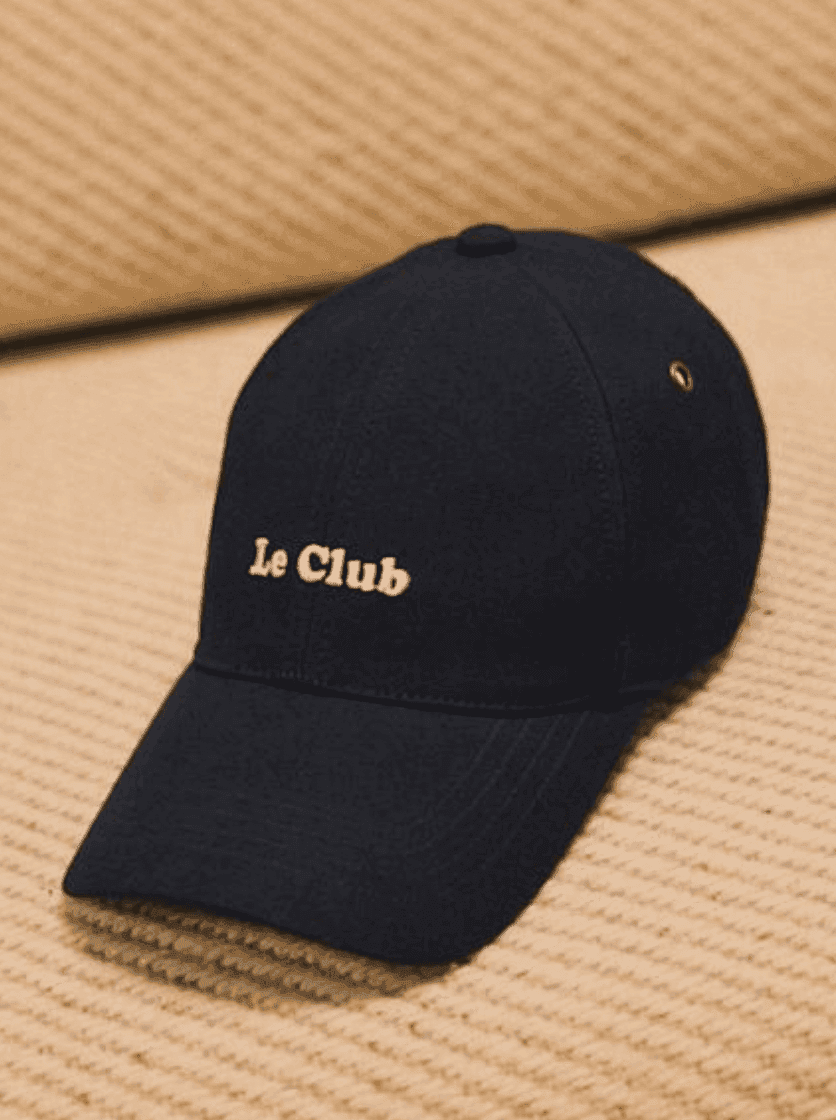 Une casquette de baseball noire avec « Le Club » brodé en blanc sur le devant, reposant sur une surface texturée beige.