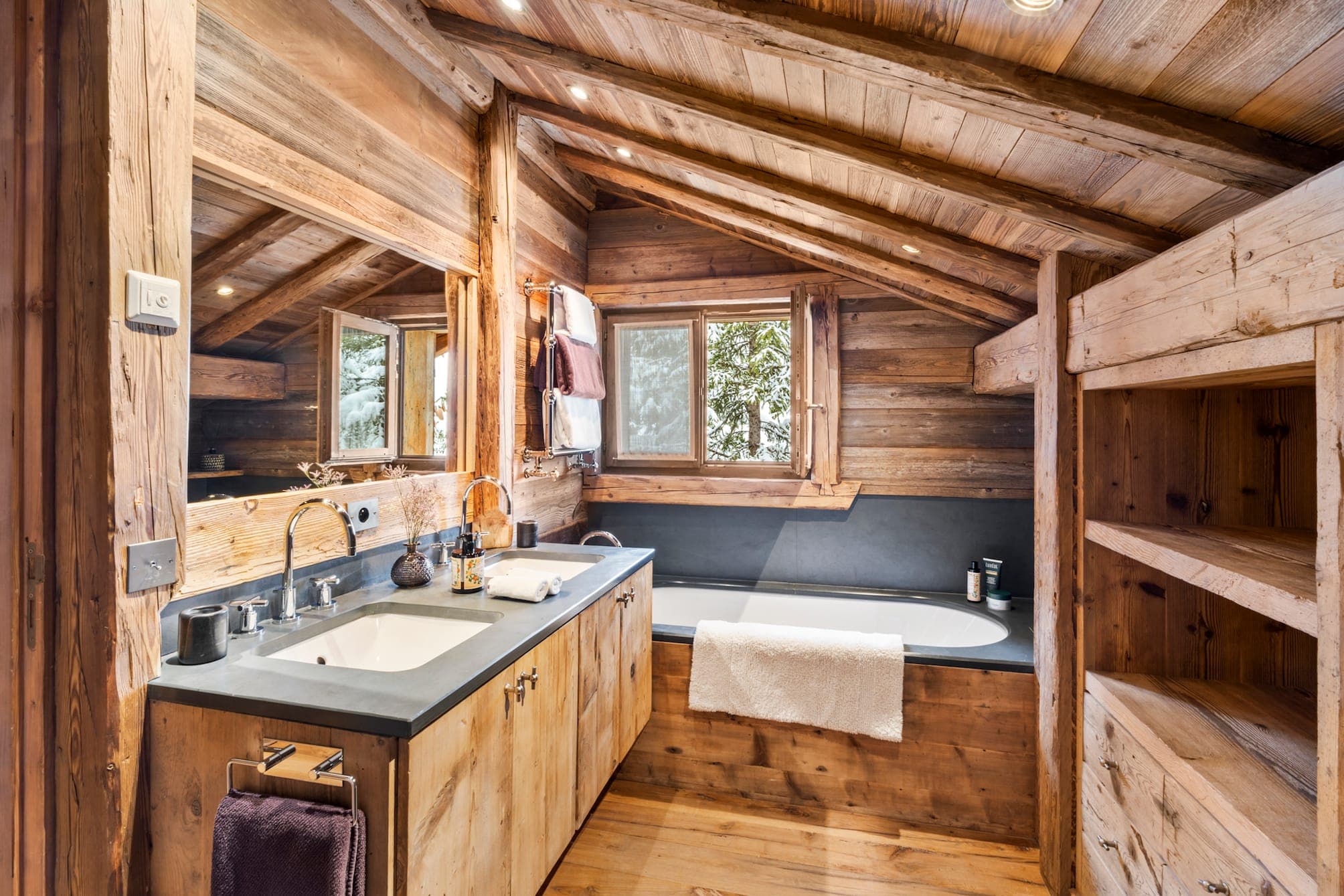 Rustic wooden bathroom with sloping ceiling, featuring a white double vanity, large mirror and comfortable bathtub with towel draped over the side. There's a window overlooking the trees and open shelving to the right.