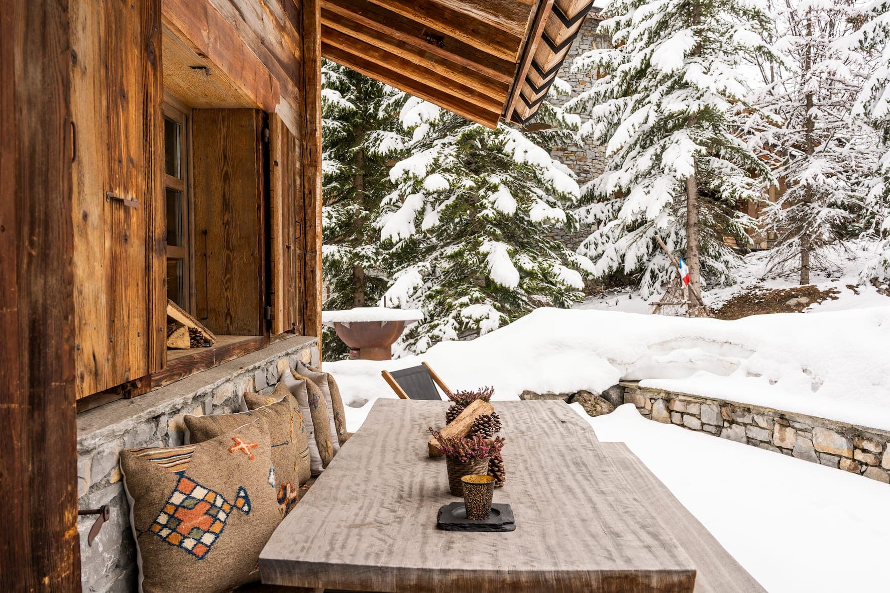 The terrace of a cozy wooden cabin, complete with table and cushions, is covered in snow. Tall pine trees and a stone wall surround the area, creating a serene winter scene.