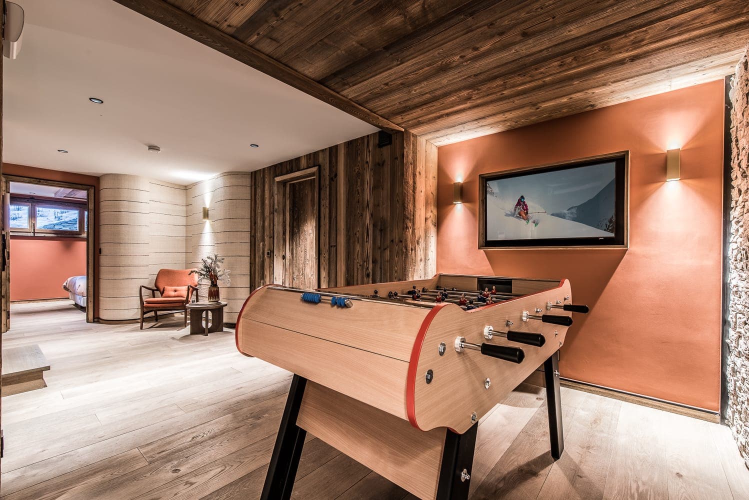 A rustic game room with wooden walls and floors. A foosball table is centrally placed under a framed ski-themed picture. An armchair and a small side table with flowers are visible near a doorway leading to a bedroom. Natural light enters through a window.