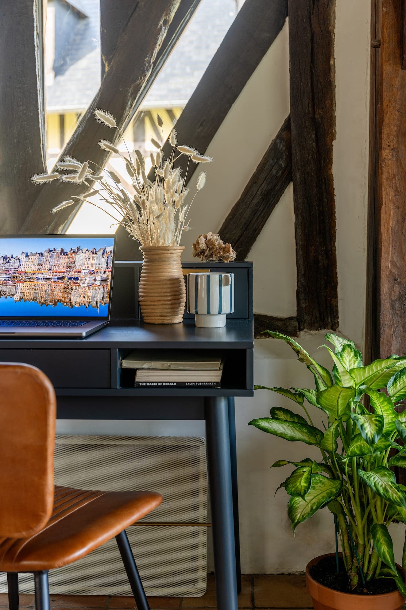 Espace de travail moderne avec un bureau noir élégant sur lequel se trouve un ordinateur portable affichant un paysage urbain. Un vase marron avec des fleurs séchées et une jardinière rayée se trouvent également sur le bureau. Une chaise en cuir marron se trouve devant et une plante verte se trouve à côté du bureau près des poutres en bois.