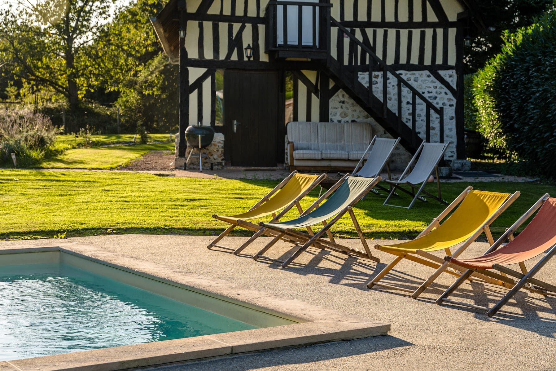 Une cour ensoleillée abrite une piscine accueillante entourée de transats aux couleurs vives. À l'arrière-plan, on aperçoit une maison rustique en noir et blanc avec un escalier en bois, ainsi qu'un canapé et un barbecue.