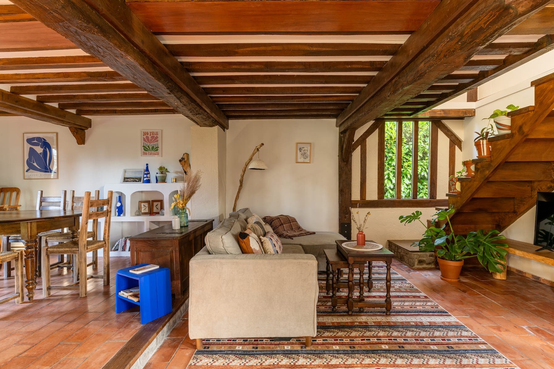 Un salon confortable avec poutres apparentes en bois, un canapé d'angle beige et un tapis rayé. Une table à manger en bois avec des chaises se trouve sur la gauche. Des escaliers avec des plantes mènent à l'étage et de grandes fenêtres laissent entrer la lumière naturelle. La décoration comprend des œuvres d'art encadrées et de la poterie.