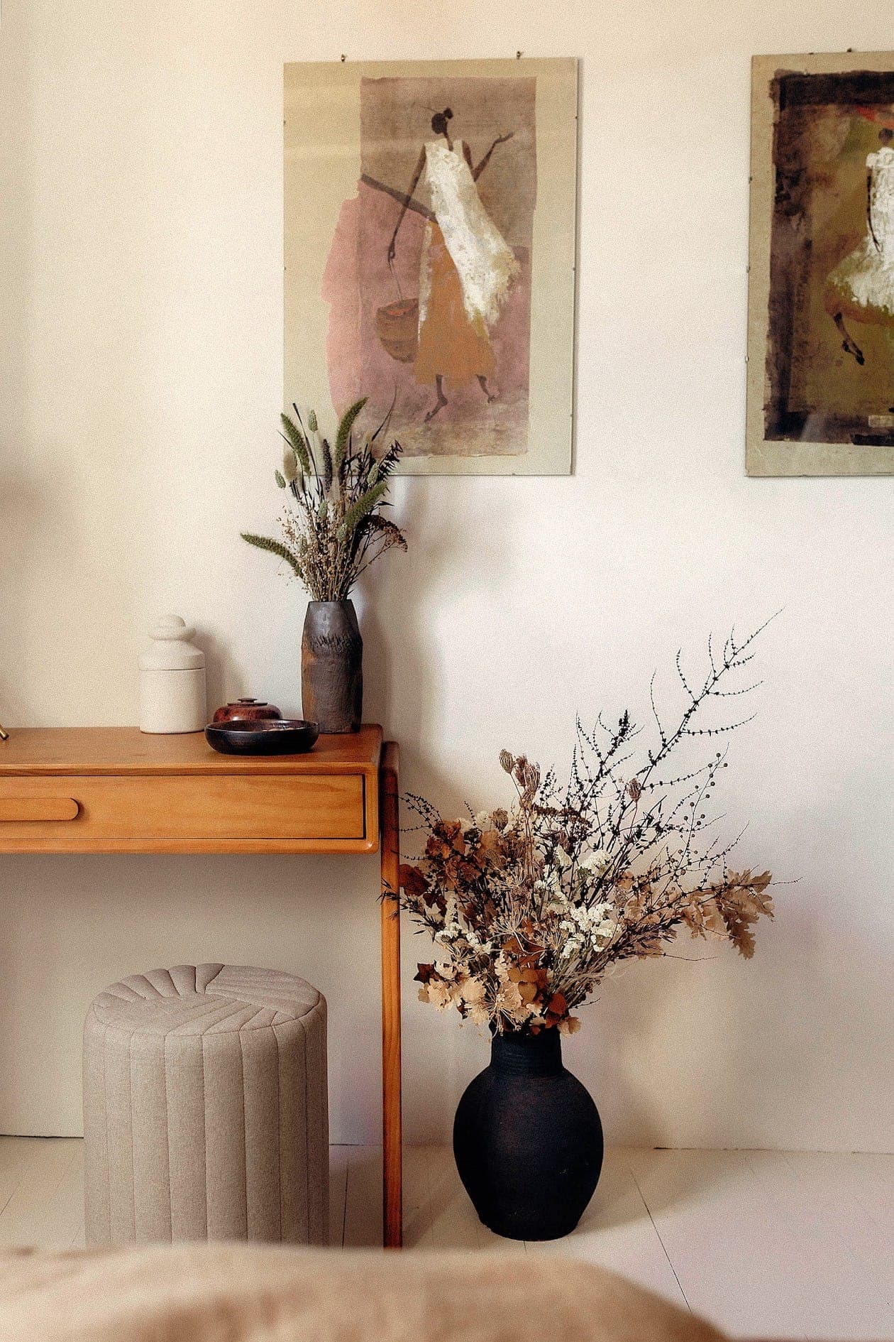 Chambre confortable avec un bureau en bois, un tabouret gris rembourré et deux tableaux abstraits encadrés sur le mur. Un vase avec des fleurs est posé sur le bureau et un autre grand vase avec des branches séchées est posé sur le sol. Les tons neutres et terreux dominent la décoration.