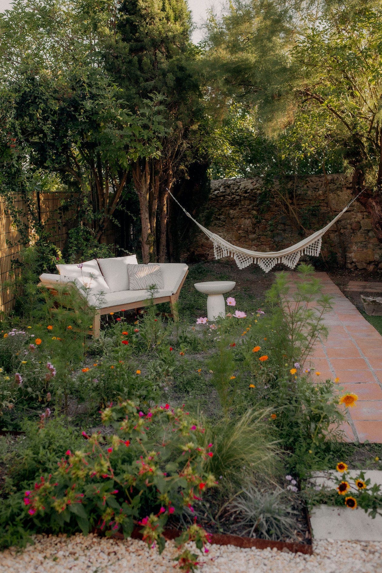 Scène de jardin avec un canapé d'extérieur beige à gauche, entouré d'une variété de fleurs colorées et de verdure. Un hamac blanc est suspendu entre deux grands arbres. À droite, un chemin de pierre traverse le jardin.