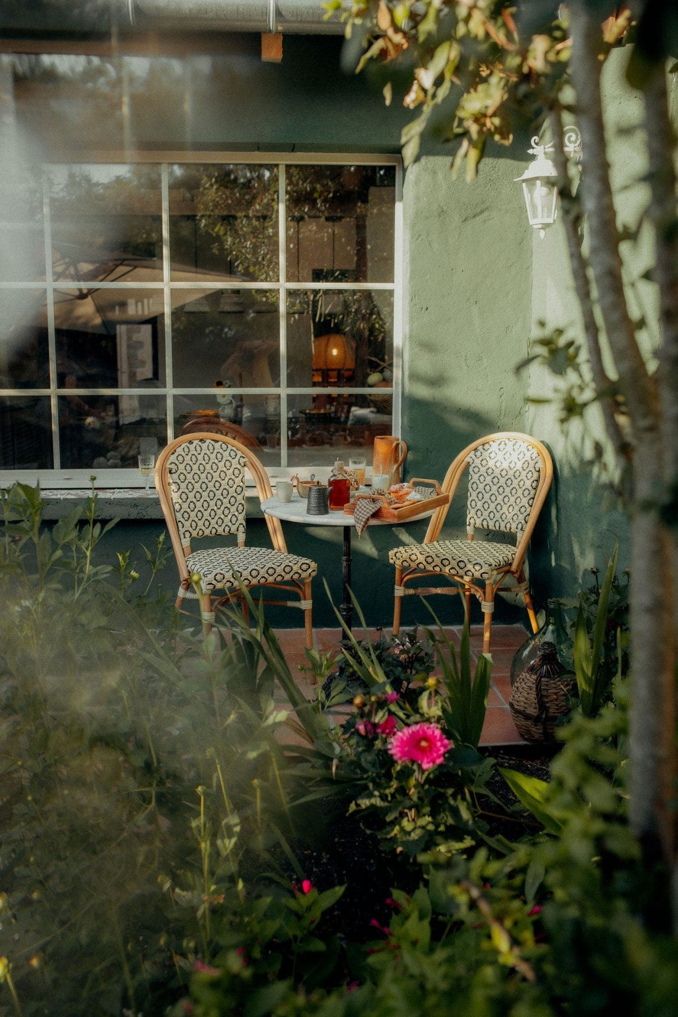 Un espace repas extérieur confortable avec une petite table ronde et deux chaises à motifs sur une terrasse. La table contient une carafe, des verres et des livres. Des plantes vertes luxuriantes et des fleurs roses entourent la zone, et une grande fenêtre se trouve à l'arrière-plan.