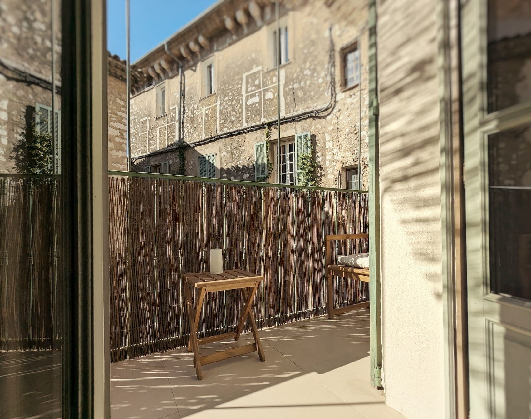 Un balcon ensoleillé avec une table et une chaise en bois, bordé d'une clôture en bambou. Le bâtiment respire le charme rustique avec ses murs en pierre et ses fenêtres à volets. Le ciel bleu vif est visible au-dessus.