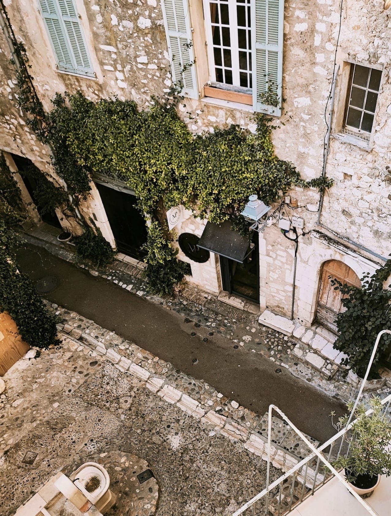 Une rue pavée étroite avec des bâtiments en pierre recouverts de lierre. La vue est d'en haut, montrant quelques petites portes et fenêtres. Des plantes en pot sont disposées le long du chemin, créant une atmosphère rustique et charmante.