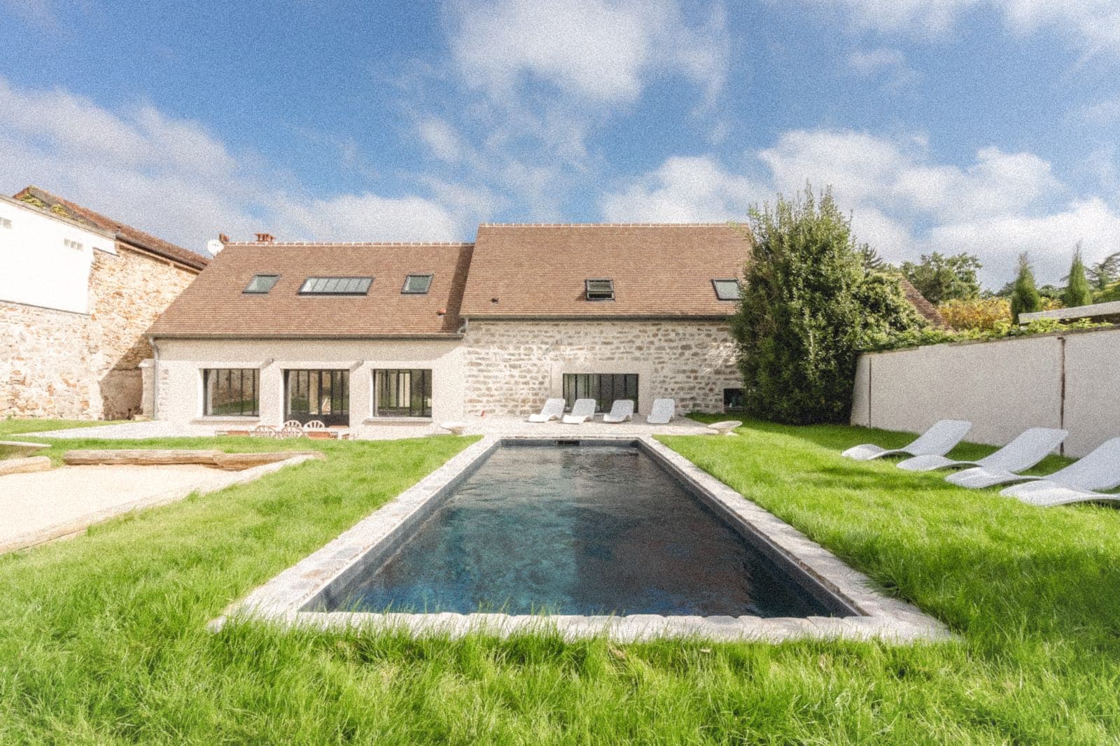 A rustic stone house with a brown roof and multiple skylights is set amidst a spacious green lawn. In front, there's a narrow rectangular swimming pool surrounded by white lounge chairs. The sky is partly cloudy.