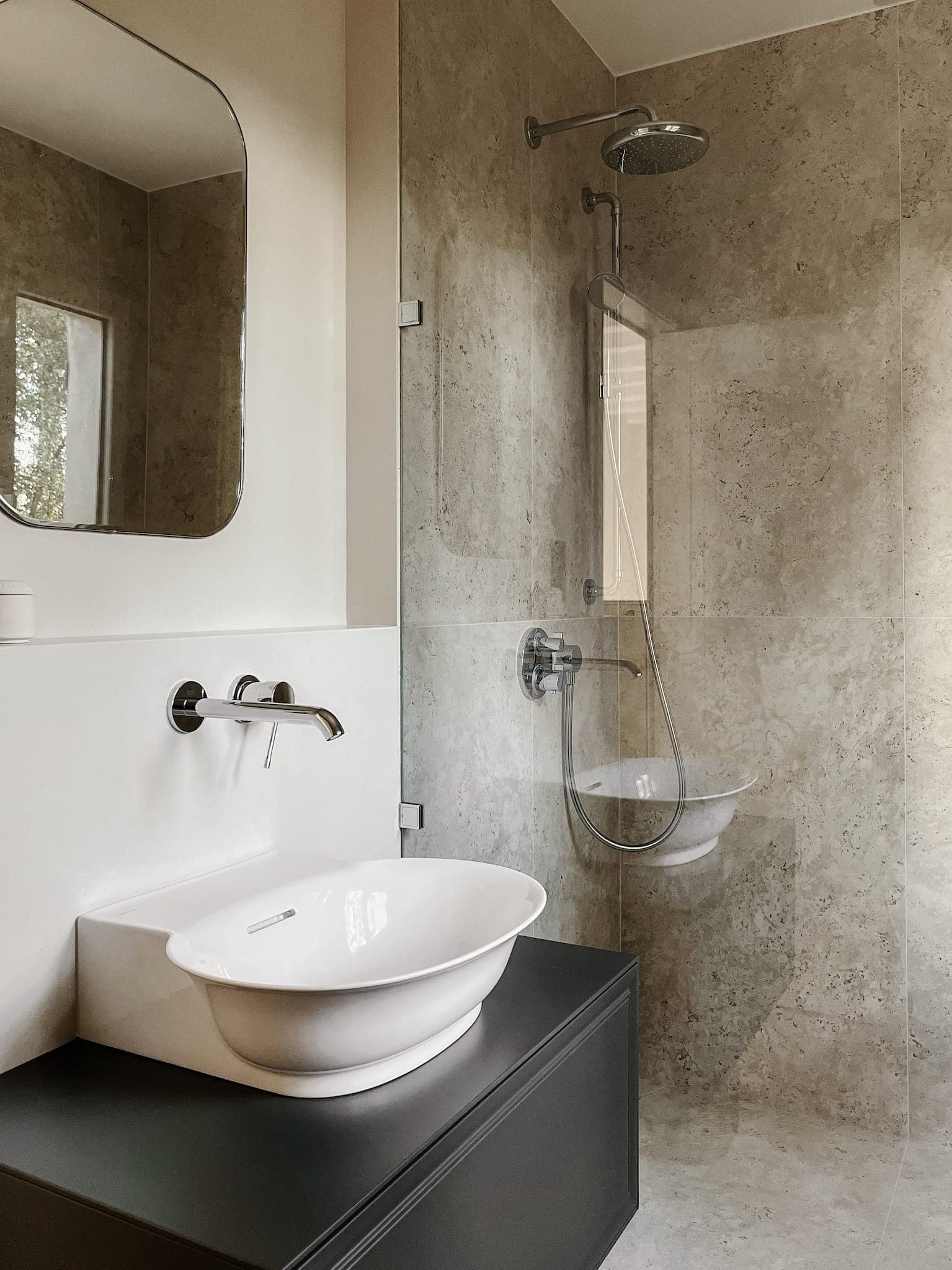 Salle de bain moderne avec miroir rond fixé au mur, lavabo élégant avec robinet intégré et douche à l'italienne avec cloison en verre et pomme de douche à effet pluie. Des carreaux de pierre beige recouvrent les murs, ajoutant une touche minimaliste et élégante.