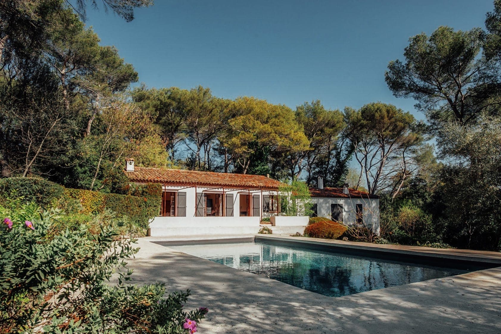 Vue d'une maison à un étage avec un toit de tuiles rouges, entourée d'arbres, par une journée ensoleillée. La maison a des murs blancs et de grandes fenêtres. Devant, il y a une piscine rectangulaire avec un reflet clair. Des plantes de jardin sont visibles tout autour.