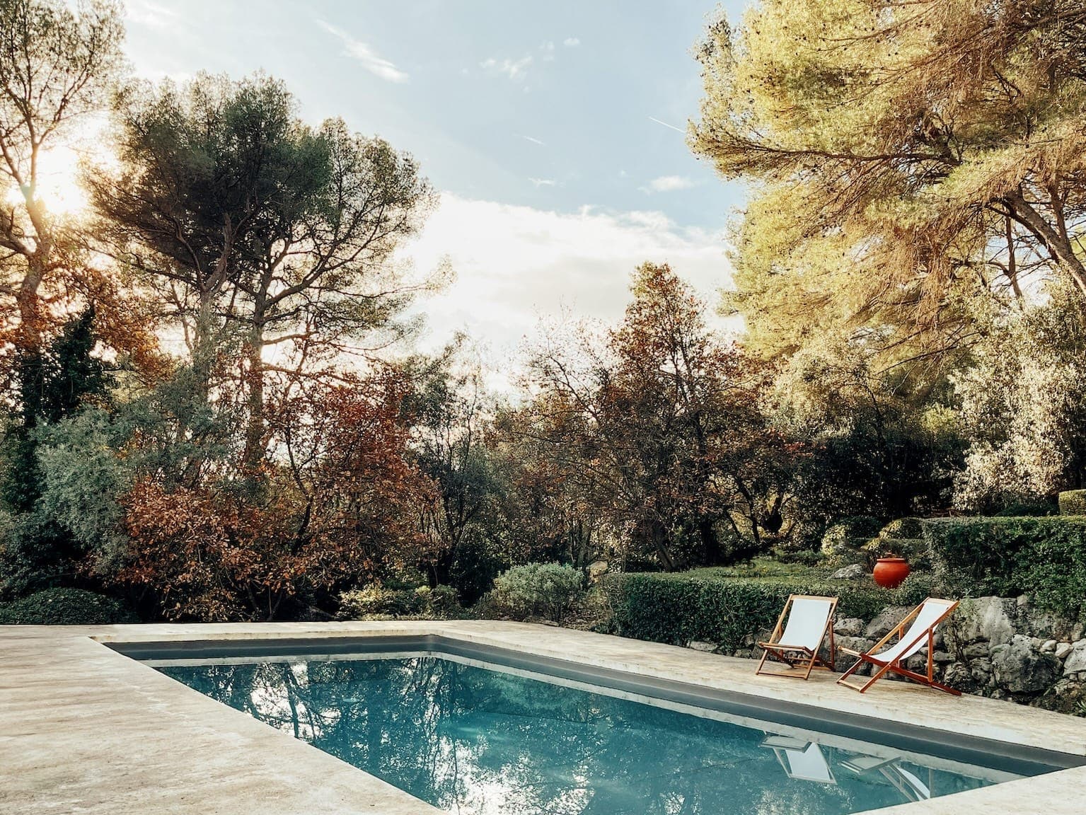 Une scène extérieure sereine avec une piscine rectangulaire entourée d'une végétation luxuriante et de grands arbres. Deux chaises longues vides sont installées à côté de la piscine sous un ciel partiellement nuageux, créant une atmosphère paisible et accueillante.