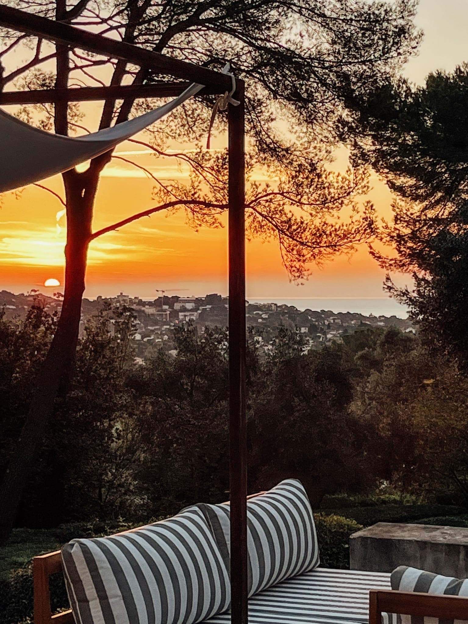 Un coucher de soleil entre les pins depuis une terrasse avec une banquette rayée.