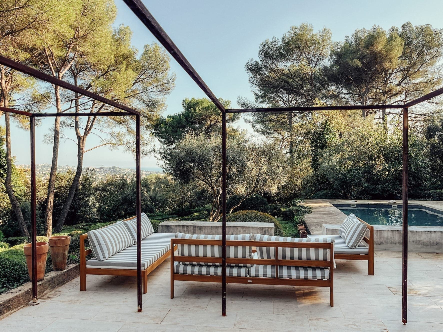 Terrasse extérieure avec banquette à coussins rayés sous une structure métallique simple. Entourée de verdure luxuriante et d'arbres, une piscine est visible en arrière-plan. Le ciel est clair, créant une atmosphère paisible et accueillante.