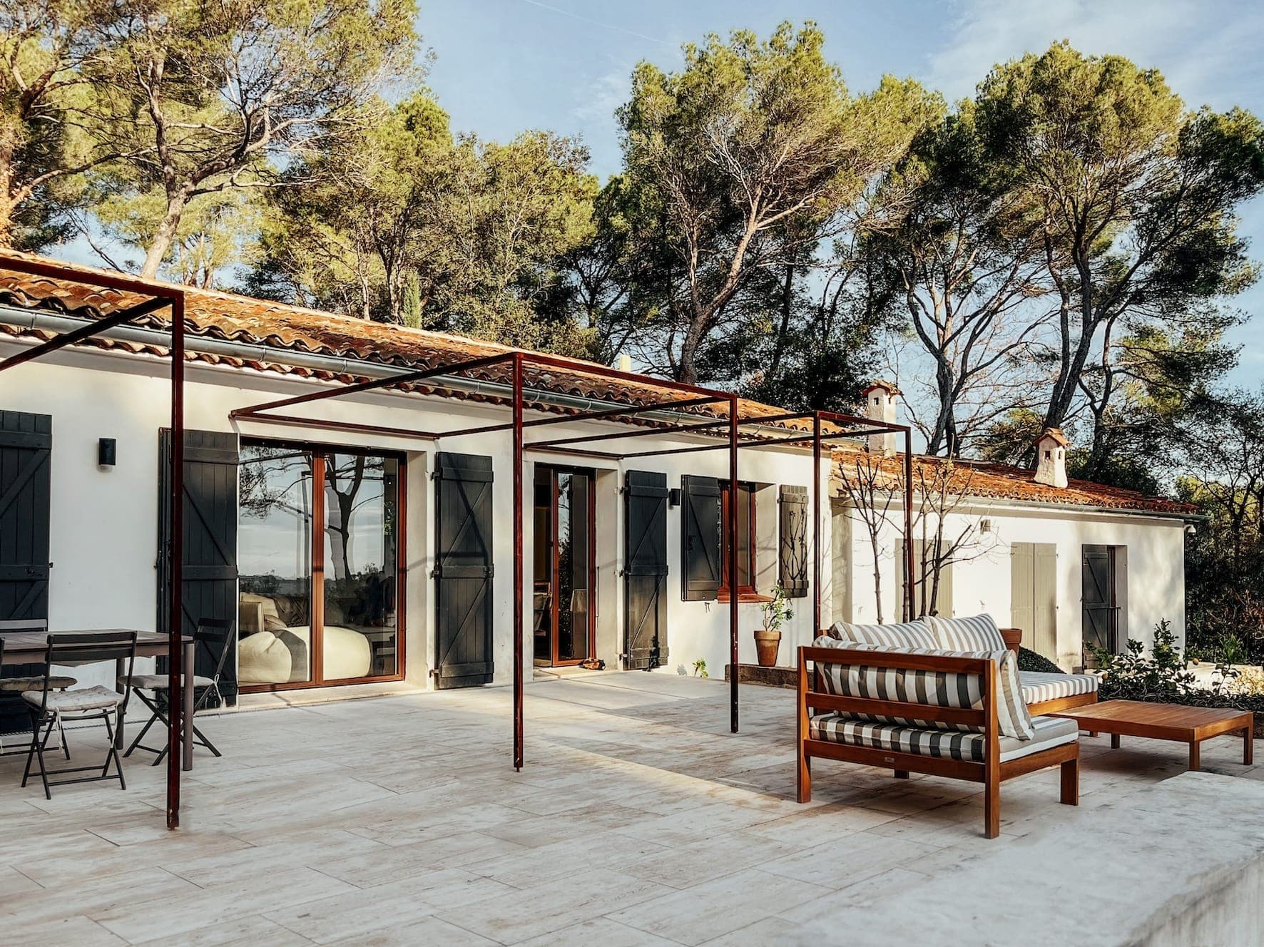 Un patio serein d'une villa blanche avec des pergolas en bois et un toit en tuiles. L'espace comprend des sièges extérieurs avec un coussin rayé, entourés de grands pins sous un ciel bleu clair, créant une atmosphère paisible et accueillante.